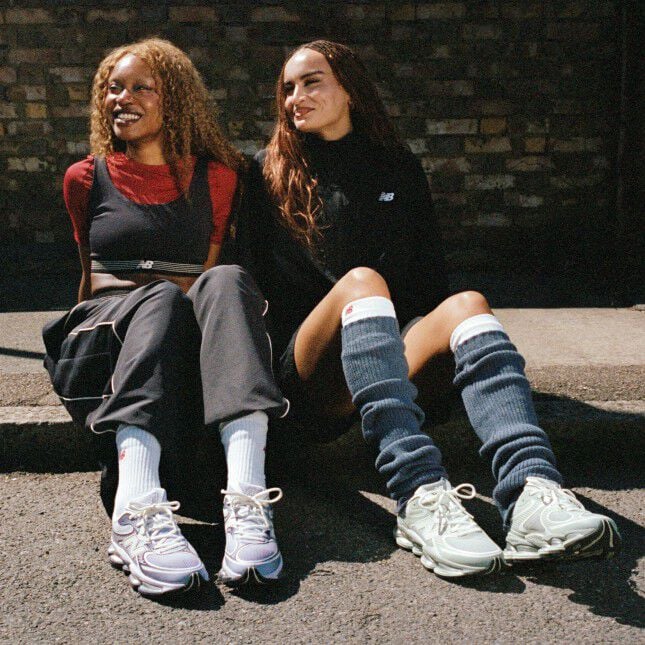 Two women seated on a sidewalk wearing New Balance sneakers.